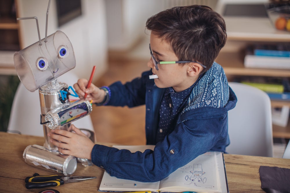 Um garoto está sentado à mesa, com uma canetinha na mão e um caderno à sua frente. Ao seu lado, está um robô de brinquedo de reciclagem. 