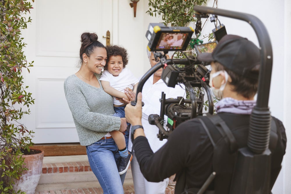 Duas mulheres estão em frente a uma casa. Uma delas segura uma criança no colo. Em frente a elas está um homem com um equipamento de filmagem.