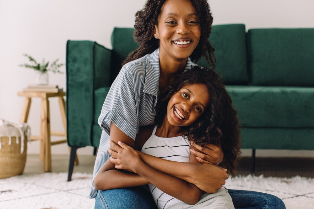 Mãe e filha abraçadas e sentadas no chão, sobre um tapete, em frente a um sofá.