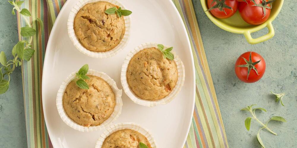 tigela branca com quatro muffins dentro. A tigela está sob uma toalha listrada. Decorando o espaço, ramos de manjericão e um bowl amarelo com tomates frescos.