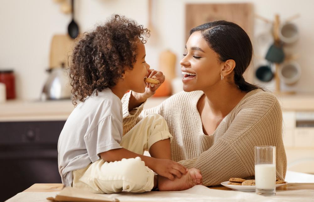 A reeducação alimentar é representada pela foto de uma mãe que oferece um biscoito ao filho. A criança está sentada em uma mesa, onde há leite e biscoitos.