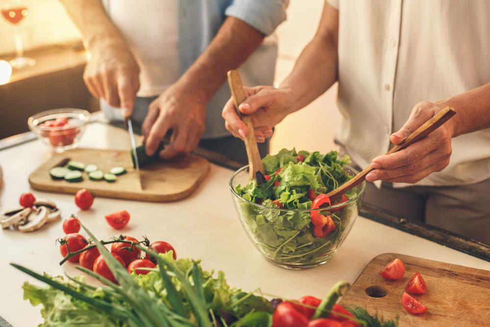 Alimentação saudável: a foto mostra a mão de dois adultos preparando uma salada com folhas verdes, pepino e tomate-cereja.