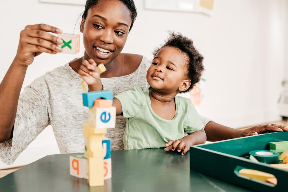 Mãe está com seu filho sentado em seu colo. Eles estão em frente a uma mesa, onde há brinquedos para bebês, e eles empilham as peças juntos. 
