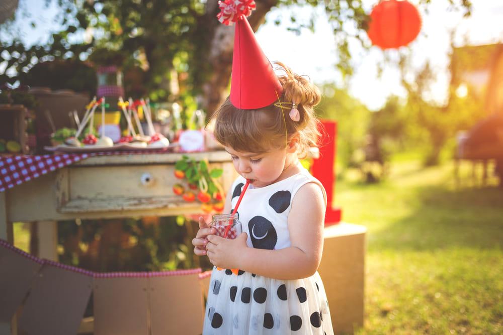 A foto mostra uma menina em uma festinha de aniversário. Ela está de vestido, chapéu de aniversário e segurando um copo com um canudinho.