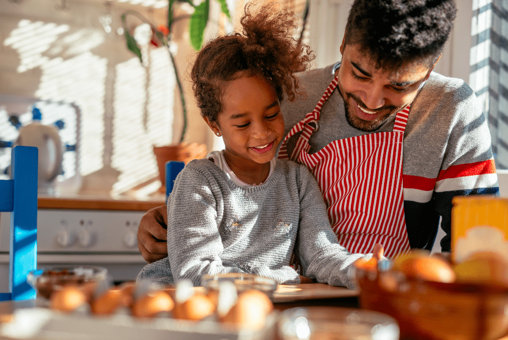 A relação de pai e filho é ilustrada por uma imagem onde a menina é abraçada pelo pai, que veste avental, enquanto eles olham para uma mesa de café da manhã com alguns alimentos.