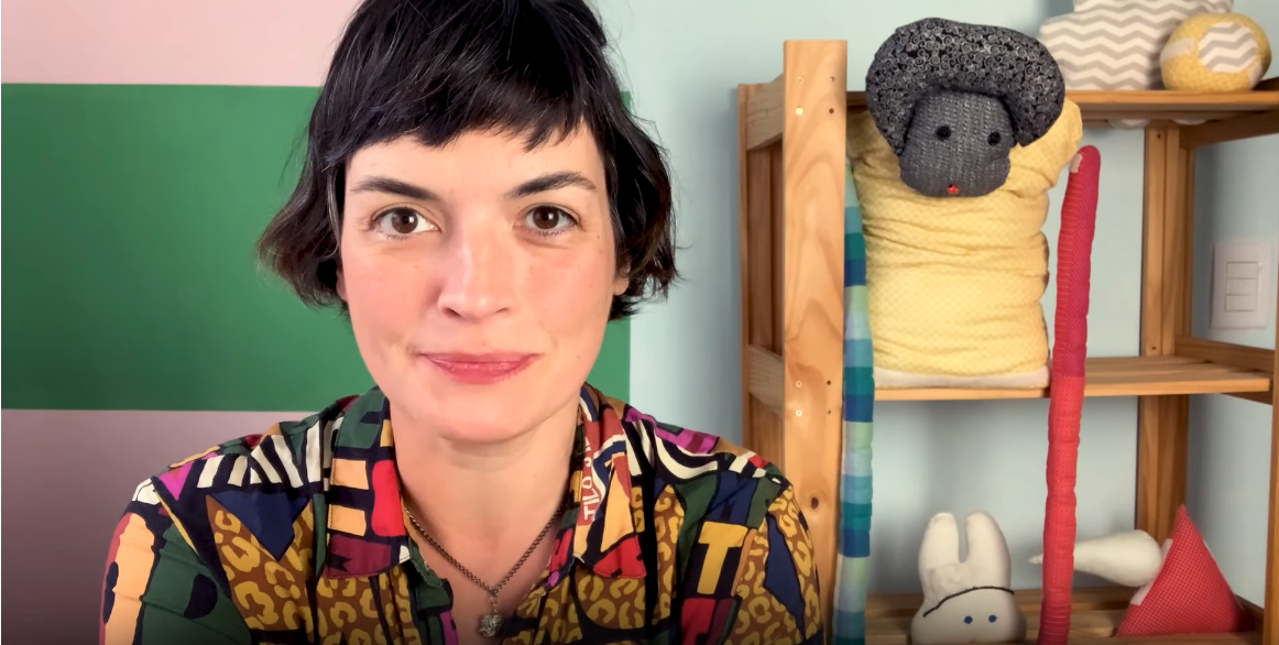 Flávia Scherner, que fala de livros infantis no canal Fafá Conta Histórias, olha para a câmera em um cenário com alguns brinquedos ao fundo.