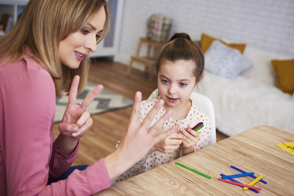 Uma mulher mostra números com os dedos, prática comum no ensino de matemática para crianças, para uma menina que a observa. 