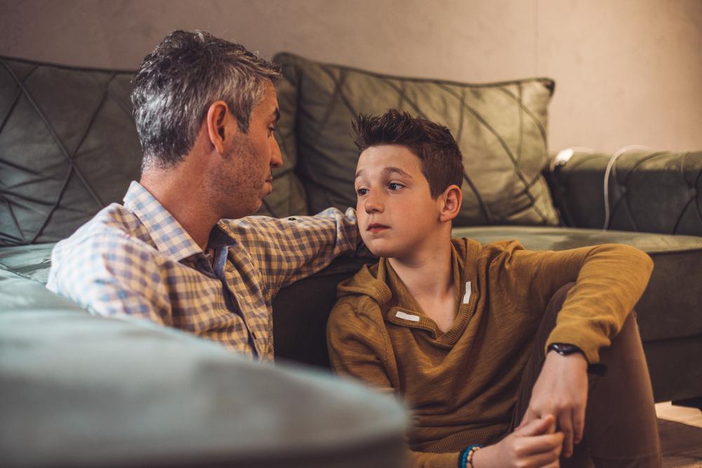 Um homem de cabelo grisalho, está sentado no chão com um menino, conversando. É uma situação em que comumente é preciso responder perguntas difíceis de crianças.