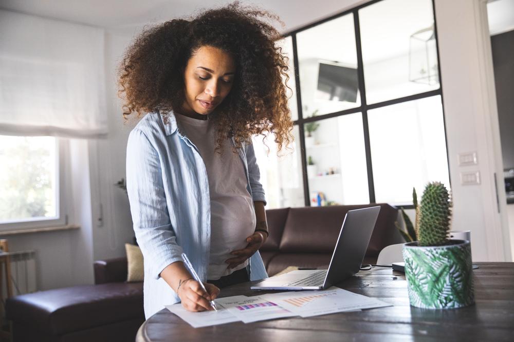 Uma mulher grávida escreve em alguns papéis na mesa da sala de casa, enquanto está de licença-maternidade. 