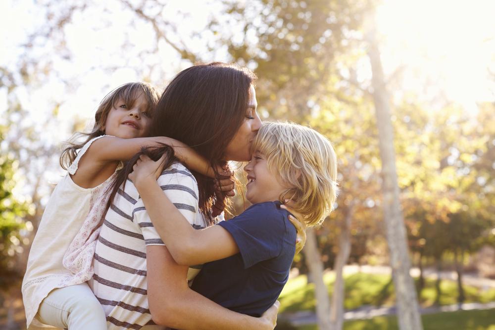 Uma mãe está junto de suas duas crianças ao ar livre. Uma está abraçada ao seu pescoço e a outra recebendo um beijo na testa