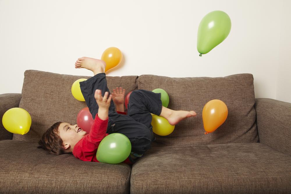 Menino deitado em um sofá com as penas para o alto brincando com bexigas coloridas. A foto faz alusão ao tema de festa de aniversário em casa. 