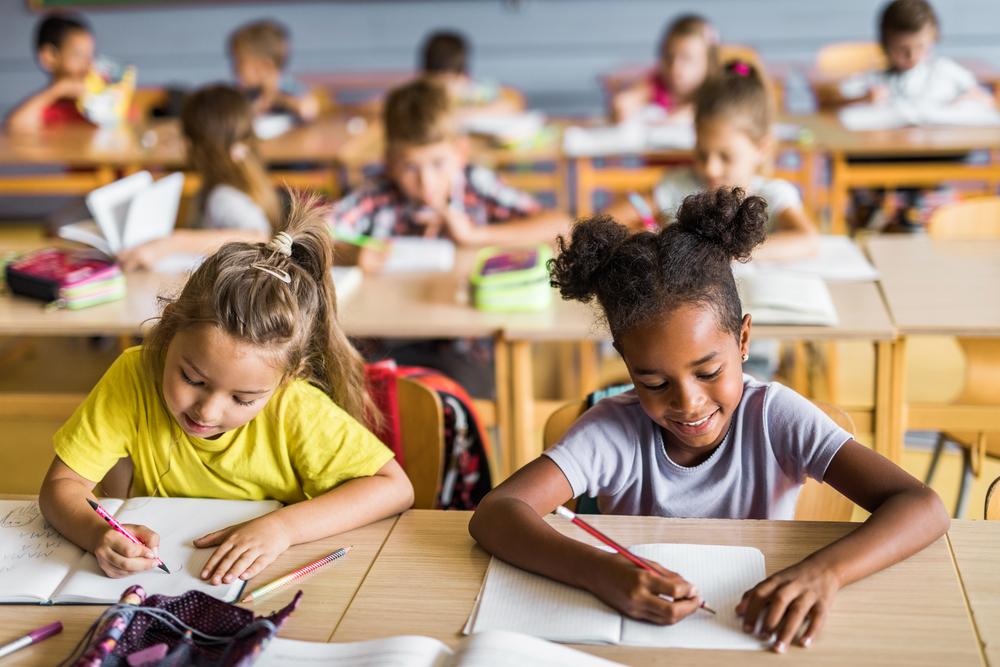 Em uma sala de aula, dez crianças estão sentadas, escrevendo sobre suas mesas de madeira. As duas meninas que estão na frente, em destaque, vestem camiseta amarela e cinza e escrevem em seus cadernos. A foto faz alusão ao tema sobre Piaget. 