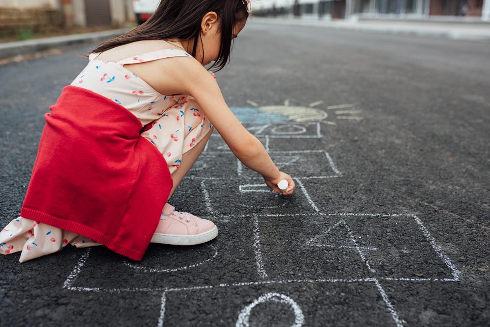 Uma menina de vestido e tênis está agachada na rua desenhando uma amarelinha no chão. A foto faz alusão ao tema de brincadeiras de rua