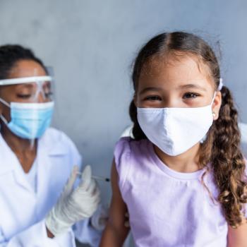 Vacinação infantil: quais vacinas meu filho precisa tomar? Um enfermeira de máscara e proteção facial realiza a vacinação infantil. Ela aplica a vacina com uma seringa em uma garotinha que está sentada e também usa máscara.