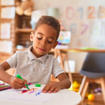 Uma criança de 4 anos está em uma sala de aula, sentado à mesa, e usa uma caneta hidrocor para desenhar. 