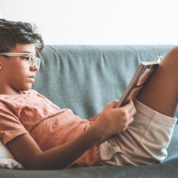 Criança de 7 anos lendo um livro deitada em um sofá, apoiada em uma almofada.