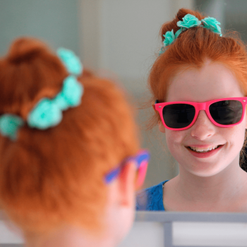 Menina sorri para a própria imagem no espelho. Se trata de uma criança de cerca de 9 anos usando óculos escuros com armação rosa.