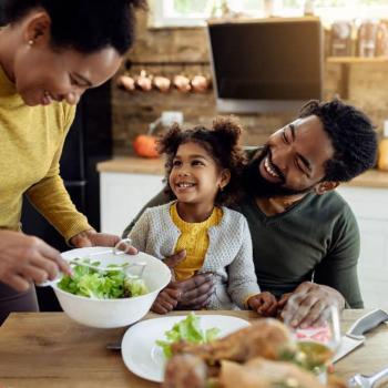 Mãe serve salada no prato para a sua filha, em busca de fortalecer a imunidade. A criança está sentada no colo do pai e todos sorriem.