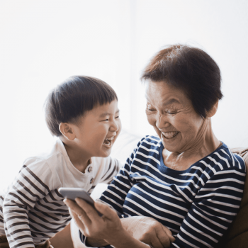 Avó e neto riem de uma brincadeira online na tela do celular, que está na mão dela.