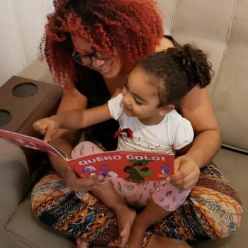 Monique Gonçalves lê livros para a sua filha. A leitura é de um livro chamado "Quero Colo!" e as duas estão sentadas no sofá.