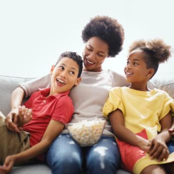 Mãe, com uma vasilha de pipoca no colo, abraça filho e filha sentados em um sofá para assistir filmes sobre maternidade. Todos sorriem.