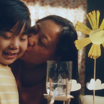 Representando a relação de mães e filhos, a foto mostra uma mãe beijando seu filho na bochecha enquanto ambos sorriem. Em frente deles, há uma flor feita de papel. 