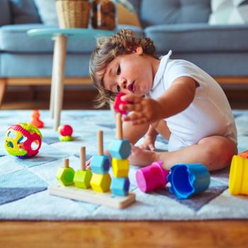 Um bebê está sentado no tapete de uma sala de estar mexendo com brinquedos de empilhar e montar, que é uma das brincadeiras que estimulam o desenvolvimento infantil. 