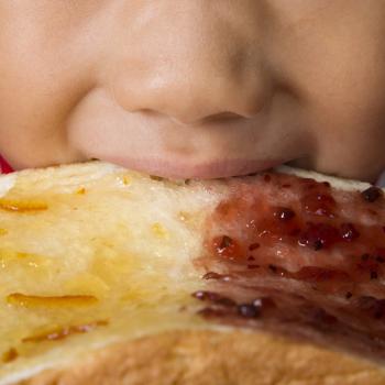 Do açúcar aos pães: os carboidratos na alimentação infantil A parte da boca e do nariz do rosto de uma criança aparece na foto. A criança morte um pão com geleia, ilustrando a presença dos carboidratos na alimentação infantil