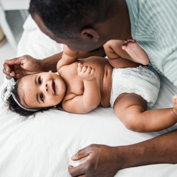 Antes do desfralde: imagem de um pai está apoiado na cama, fazendo um bebê de fralda sorrir. Ele faz carinho na cabeça da criança.  