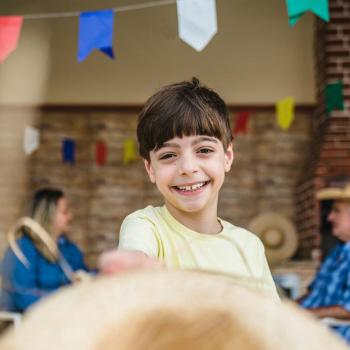 Festa junina: 5 brincadeiras típicas para fazer em casa Criança sorrindo para a foto. Ao fundo, dois adultos conversam num cenário de festa junina, com bandeirinhas coloridas penduradas.