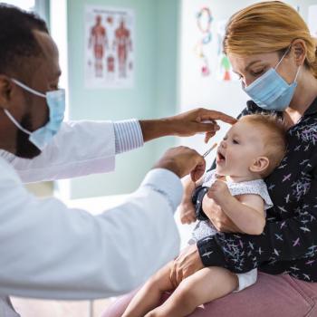 Com que frequência devemos consultar o pediatra? Em um consultório, um médico pediatra examina um bebê que está no colo de sua mãe.