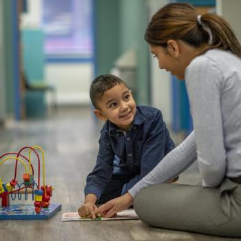 Identidade de gênero: imagem de uma criança sentada ao chão junto com a pediatra. Ambos sorriem e brincam com brinquedos que também estão no chão. 