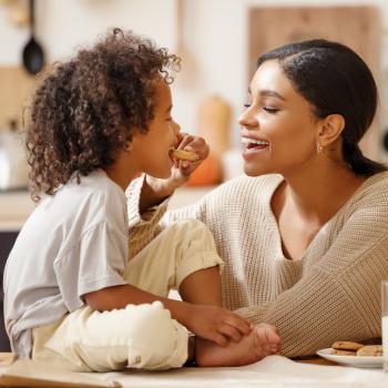 Reeducação alimentar: uma oportunidade para a família toda A reeducação alimentar é representada pela foto de uma mãe que oferece um biscoito ao filho. A criança está sentada em uma mesa, onde há leite e biscoitos.