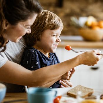 A seletividade alimentar é ilustrada pela foto de uma mãe tentando dar um tomate com um garfo para a sua criança, que está sentada em seu colo. A criança está de olhos e boca fechada, se negando a comer. 