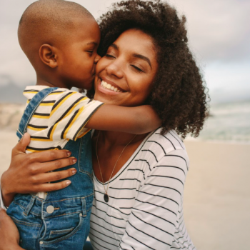 Uma mãe está ajoelhada na areia e recebe um abraço e um beijo de seu filho. Ela está com os braços ao redor dele, sorrindo de olhos fechados, e representa um dos diferentes tipos de mãe na relação com os filhos. 