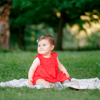 Criança de um ano com vestido vermelho e fita no cabelo posa para foto em um parque, sentado em um pequeno cobertor colocado sobre a grama.