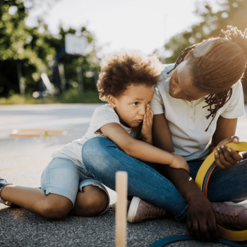 Criança de 6 anos: transições importantes estão vindo Mãe e filho, uma criança de 6 anos, estão sentados no chão de um espaço ao ar livre. A mãe olha para o filho, que o abraça.