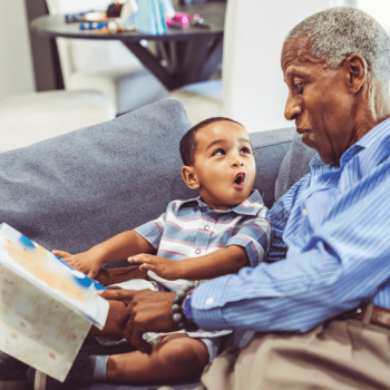Avô e neto se olham com expressão de impressionados enquanto leem livros infantis juntos, sentados no sofá.