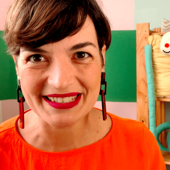 Flávia Scherner, que fala de livros infantis no canal Fafá Conta Histórias, sorri para a câmera em um cenário com alguns brinquedos ao fundo.