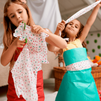 Duas crianças de cerca de 8 anos cortam papel colorido com uma tesoura sem ponta customizando pequenas peças de roupa com este material. Uma delas está utilizando, sobre a sua roupa comum, um vestido feito de papel.