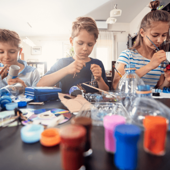 Três pré-adolescentes pintam brinquedos não estruturados sentados à mesa em uma cozinha. Esses brinquedos estão sendo criados a partir de garrafas pet cortadas, papelão e outros materiais.