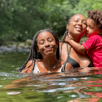 Dentro de um lago, a cantora Negra Li, ilustra a importância da música para crianças. Ela está segurando um filho no colo e em frente está sua outra filha.
