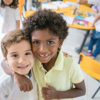 9 dicas de como criar crianças antirracistas para incluir na rotina A foto mostra dois meninos de cores e aparências distintas sorrindo, se abraçando e olhando para a câmera, dentro de uma sala de aula.