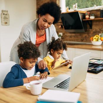 Em um ambiente de casa, uma mãe auxilia seu filho e sua filha em atividades de aulas remotas.