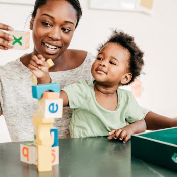 Mãe está com seu filho sentado em seu colo. Eles estão em frente a uma mesa, onde há brinquedos para bebês, e eles empilham as peças juntos. 