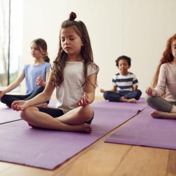 A imagem mostra quatro crianças sentadas praticando meditação infantil, com as pernas cruzadas e os olhos fechados.