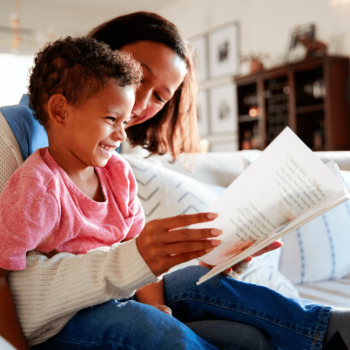 Mãe sorri com o filho sentado em seu colo enquanto os dois leem um livro juntos representando a ideia da mãe suficientemente boa.