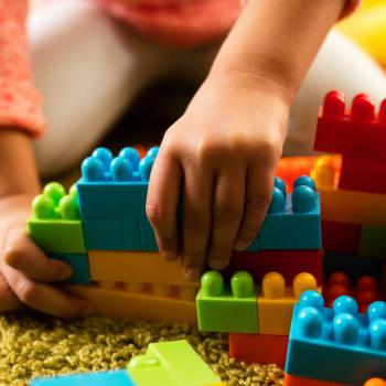 A imagem mostra as mãos de uma criança montando peças de brinquedo em cima de um tapete, representando a importância do brincar