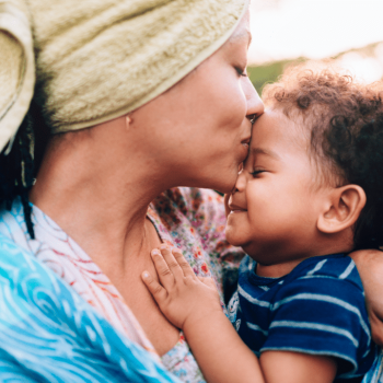 Mãe tem uma toalha de banho enrolada no cabelo e beija a testa do filho, eles estão de olhos fechados, como se satisfeitos, ilustrando a importância da rede de apoio.