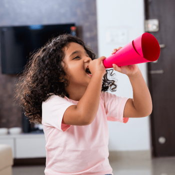 O que é TDAH infantil/hiperatividade? Saiba como identificar e lidar Menina sorri enquanto grita algo utilizando um megafone feito de papel enrolado, ilustrando a hiperatividade.